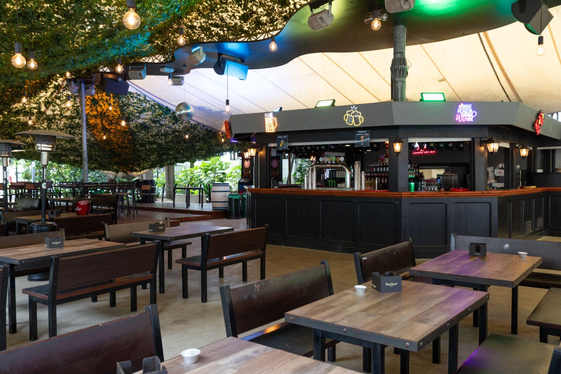 Bancone bar di La Serra Sport & Lounge con tavoli, panche in legno, luci sospese e copertura verde al Camping Mediterraneo.