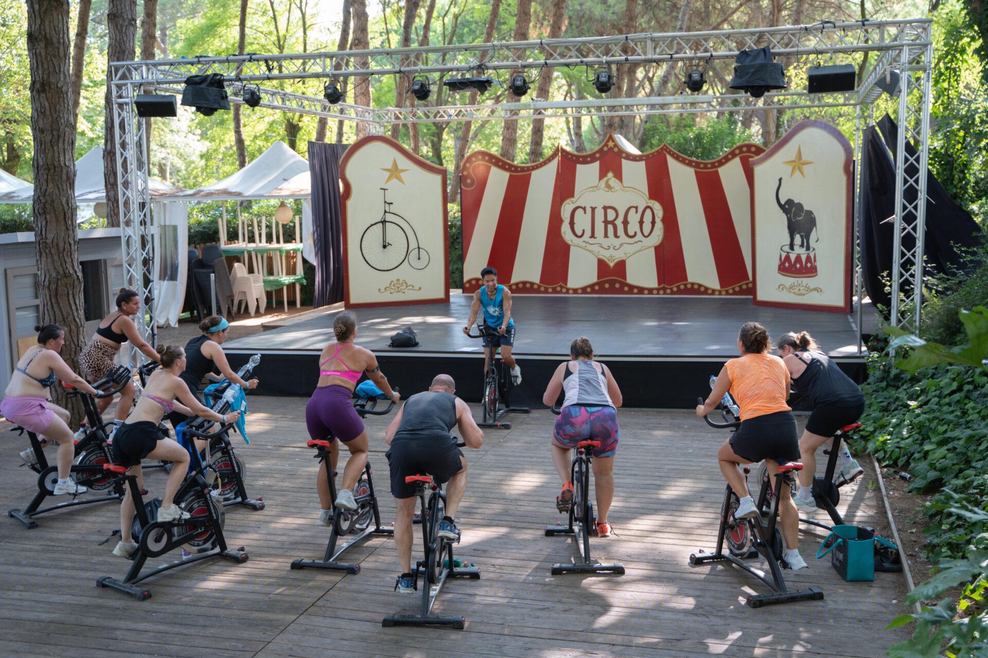 Adulti si allenano sulle cyclette durante una lezione di spinning all'aperto tra i pini del Camping Village Mediterraneo