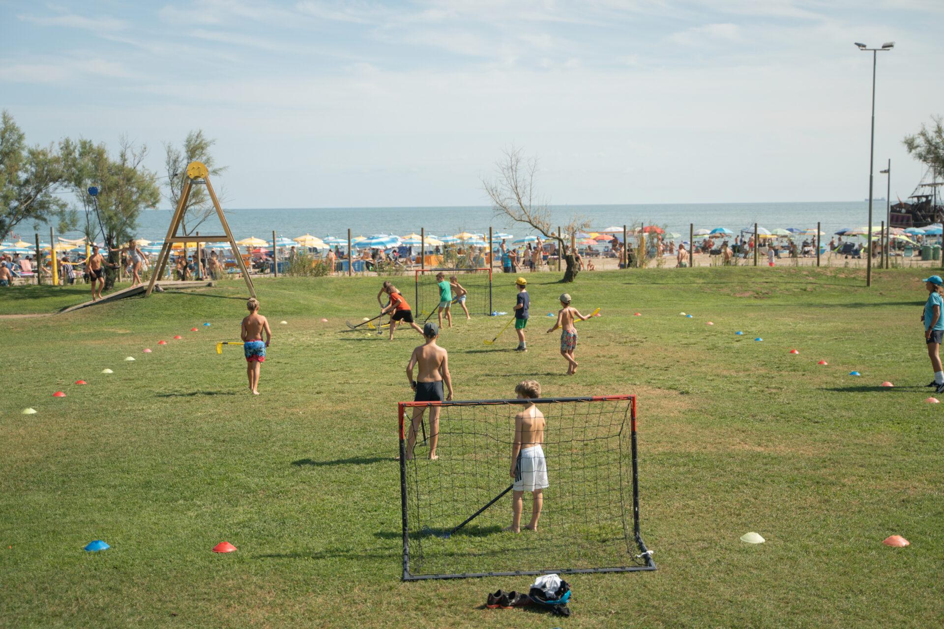 Bambini e ragazzi giocano a hockey su un prato con piccole porte, mentre sullo sfondo si vede la spiaggia del Camping Mediterraneo