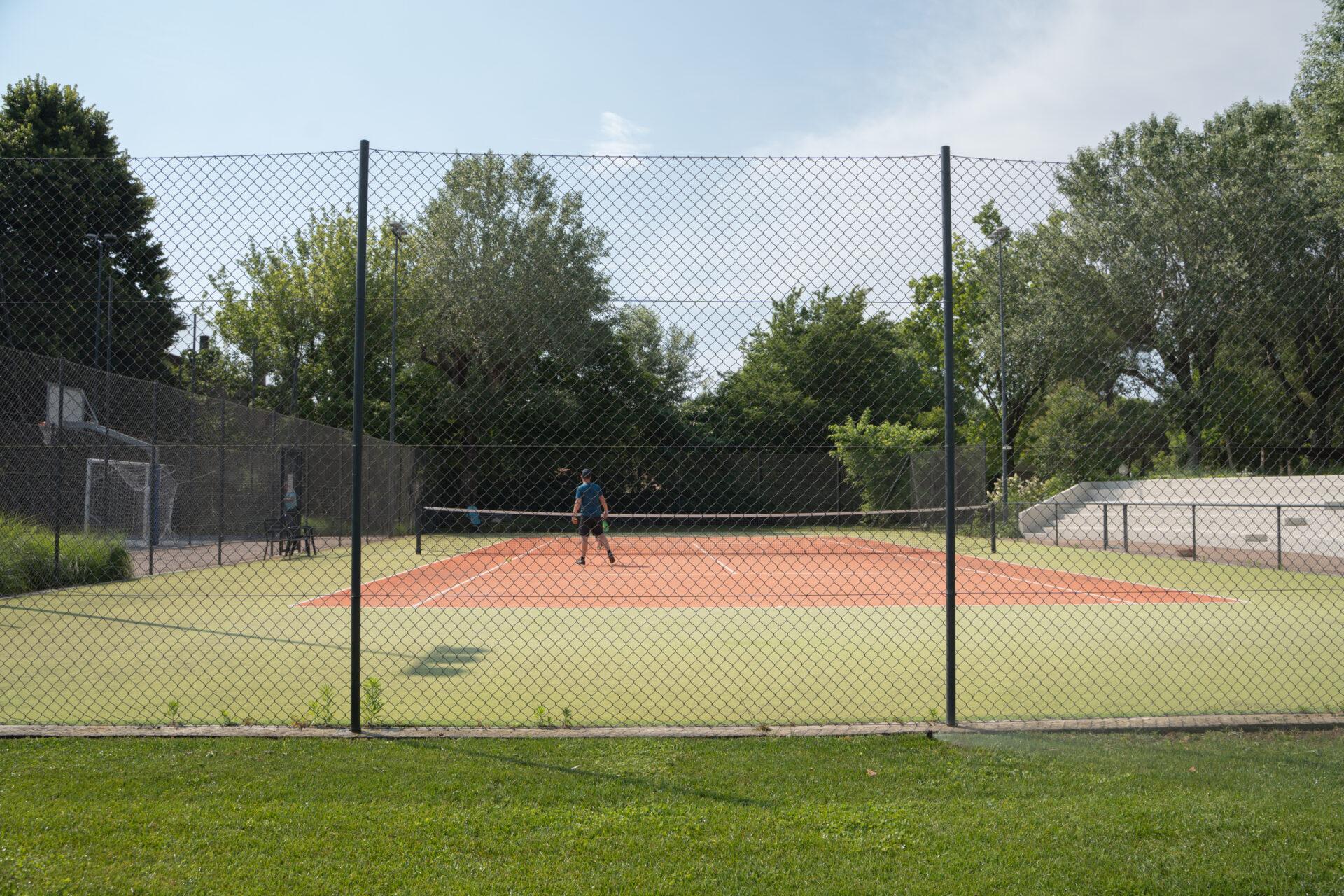 Giocatore impegnato in una partita sul campo da tennis de La Serra Sport, circondato da alberi e recinzioni