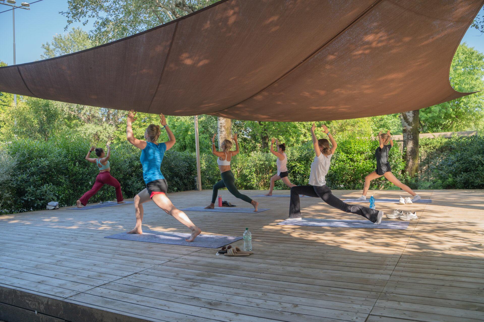 Gruppo di donne pratica yoga sui tappetini sotto una copertura ombreggiante immersa nel verde del Camping Mediterraneo