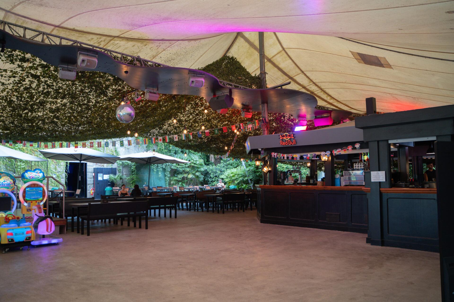 Area bar di La Serra Sport & Lounge con tavoli, ombrelloni, luci colorate, bandierine e giochi arcade.