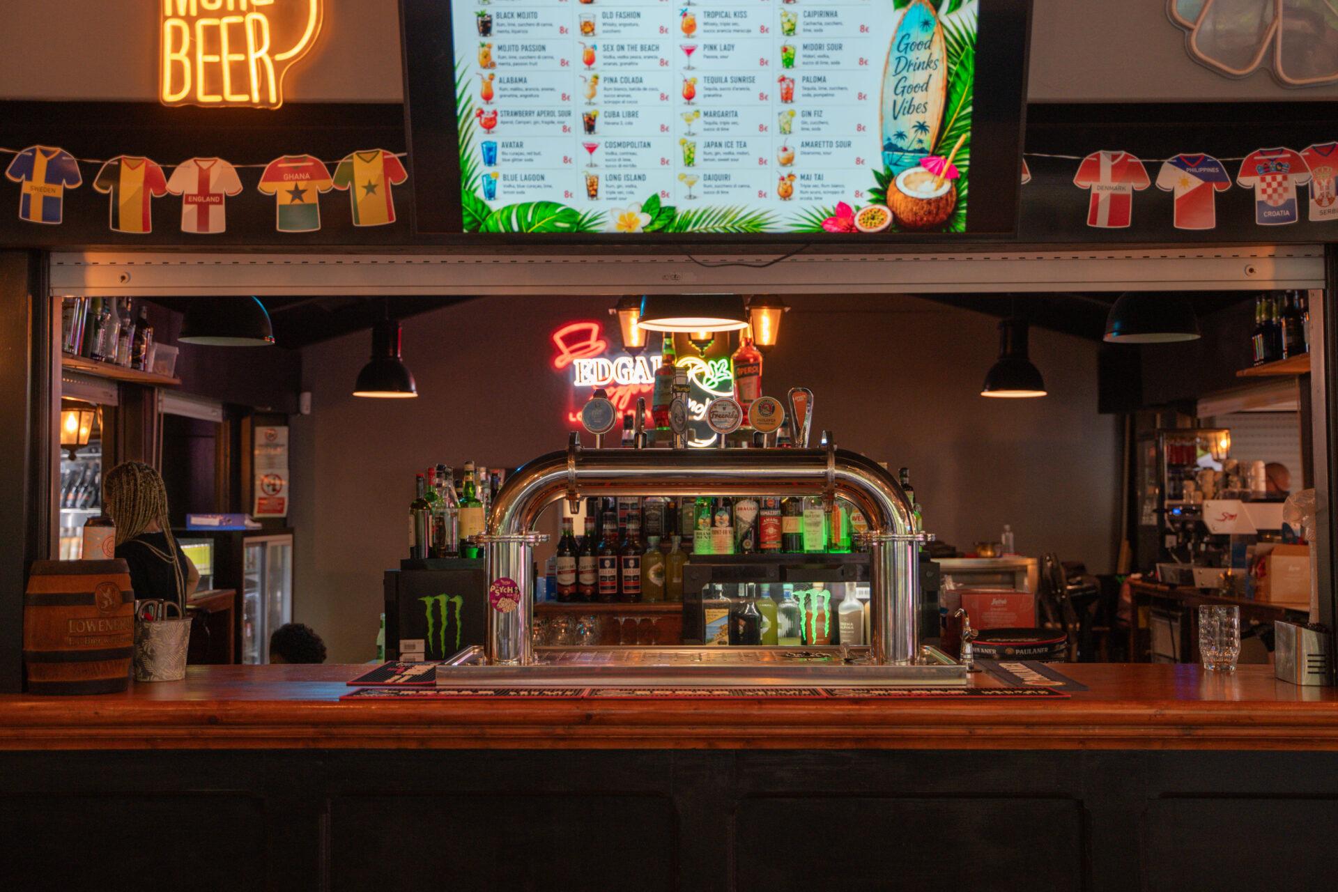 Bancone di La Serra Sport & Lounge con spine per la birra, bottiglie, insegne luminose e menù dei cocktail.