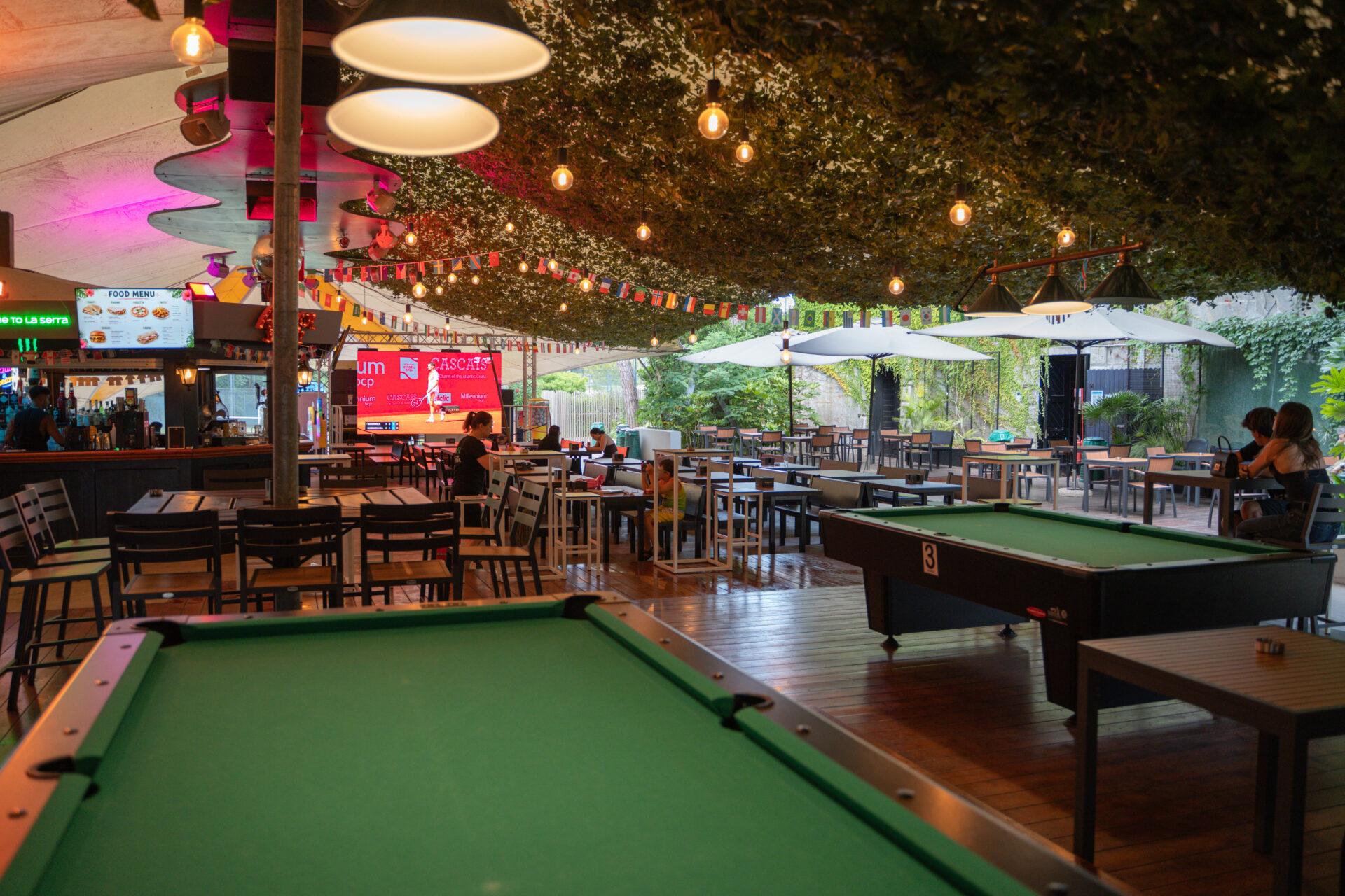Tavoli da biliardo, area bar e ospiti seduti a La Serra Sport & Lounge del Camping Mediterraneo, sotto luci sospese.